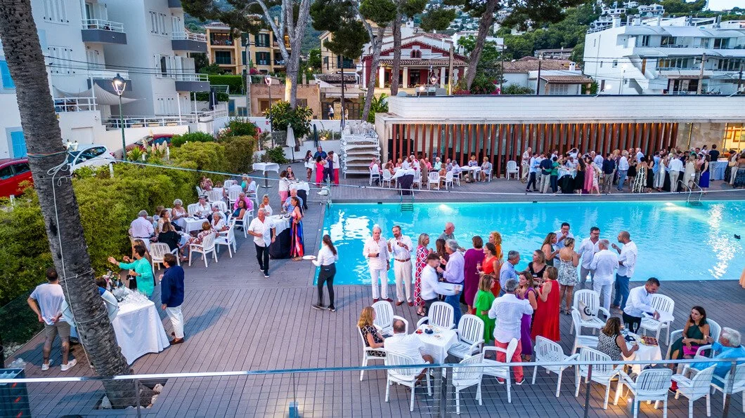 Fiesta en una piscina con muchas personas sociales, mesas con comida y bebida, en un entorno al aire libre rodeado de edificios y naturaleza.