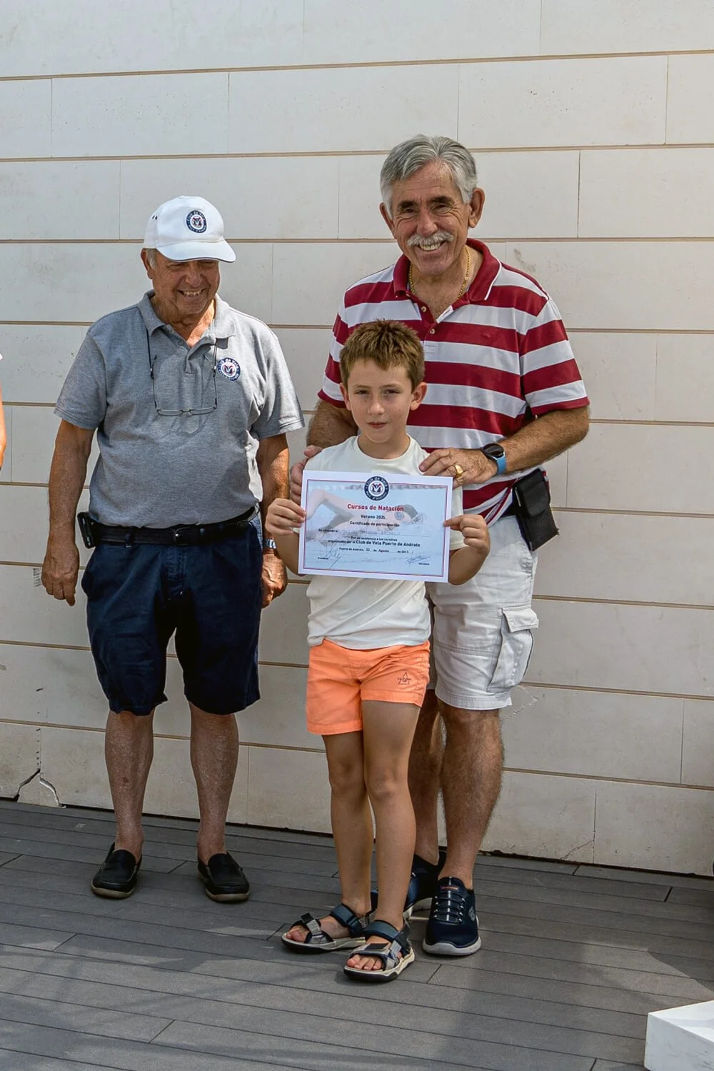 Niño sosteniendo un certificado de natación, acompañado de dos adultos, uno con camiseta a rayas y otro con gorra y camiseta gris, en un evento de premiación.