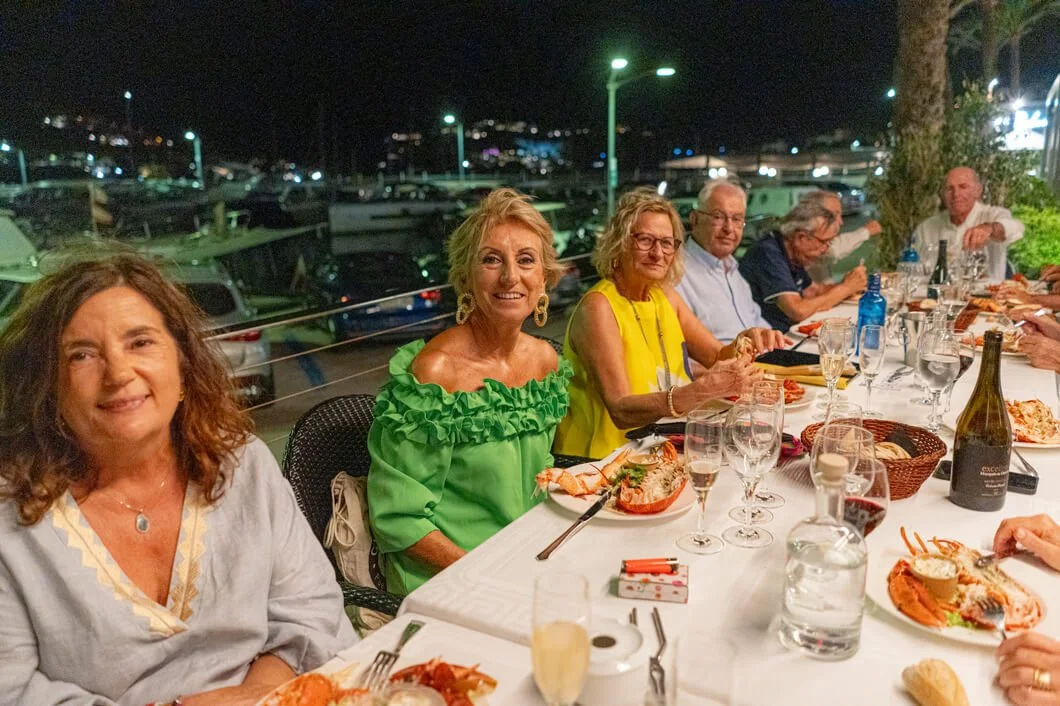 Grupo de personas cenando al aire libre en la noche, con mar en el fondo y barcos en un puerto.