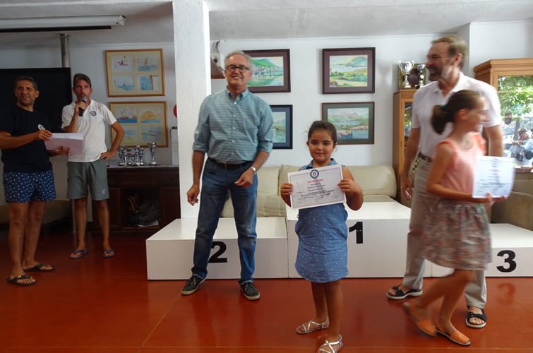 Niña en el primer lugar del podio recibiendo un certificado durante una ceremonia de premiación, con varias personas a su alrededor en un ambiente interior.