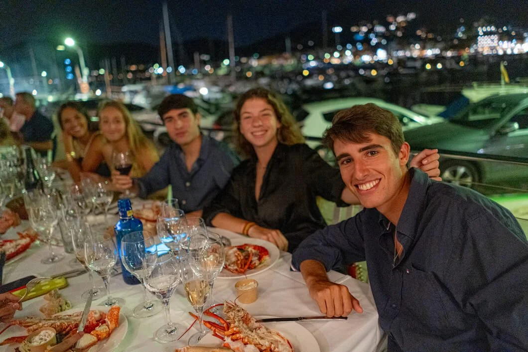 Grupo de personas disfrutando una cena en un restaurante cerca del mar, con mesas con comida y bebidas, y un fondo de yates y luces de la ciudad por la noche.
