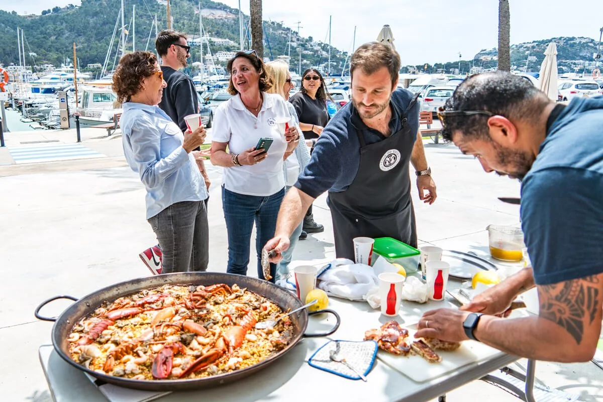 Grupo de personas en un muelle de pescado, algunos conversando y otros sirviendo paella en una mesa, con yates y montañas de fondo.