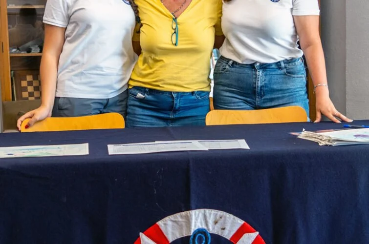 Tres personas de pie detrás de una mesa con una bandera azul, roja y blanca, en un entorno interior.