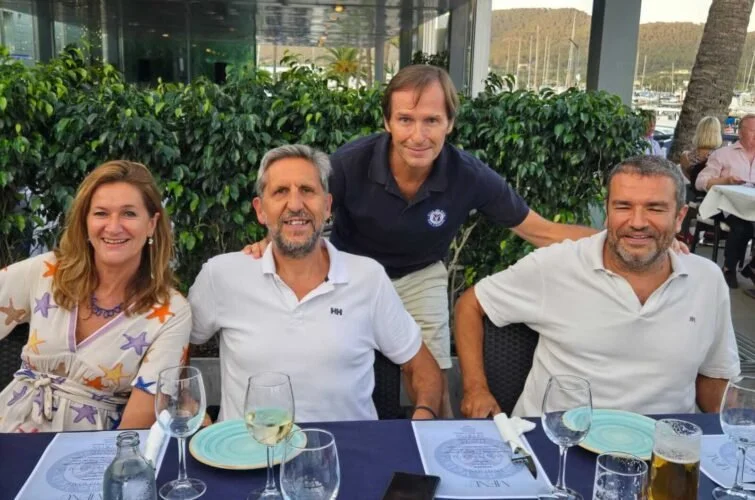 Cuatro personas sentadas y una más de pie en un restaurante, sonriendo, con mesas y vasos en primer plano, en un ambiente al aire libre con vegetación y vista a un puerto