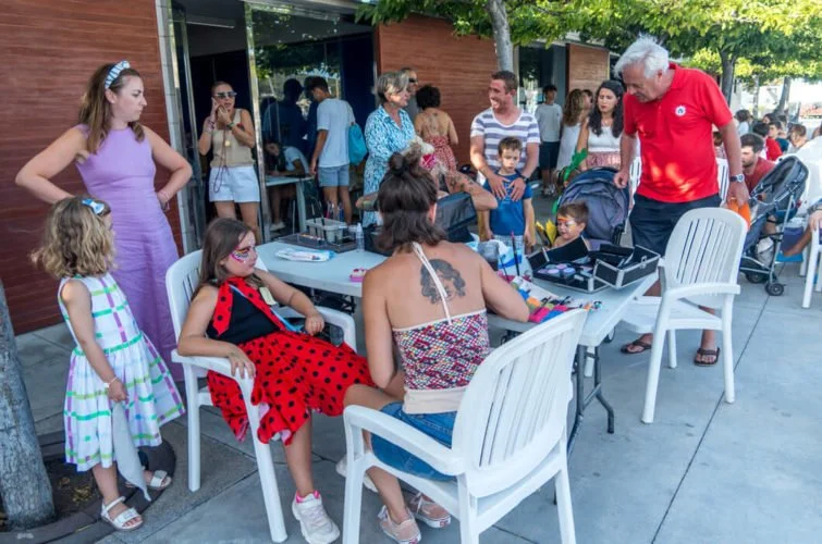 Personas, incluyendo niños, en una calle, participando en una actividad artística o manual, con algunas personas observando y otras interactuando con la artista.
