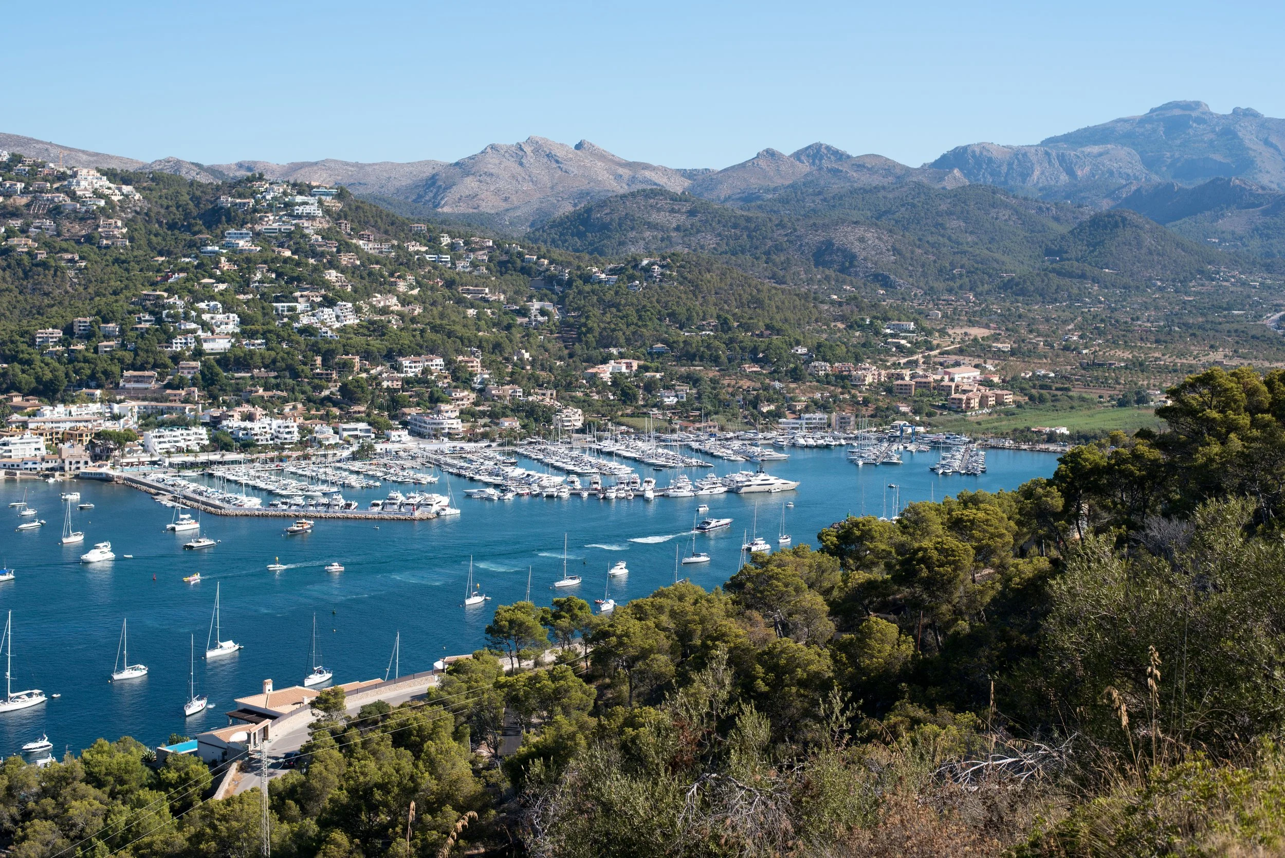 Vista panorámica de un puerto con yates y barcos, rodeado de colinas con casas y vegetación, y montañas en el fondo bajo un cielo claro y despejado.