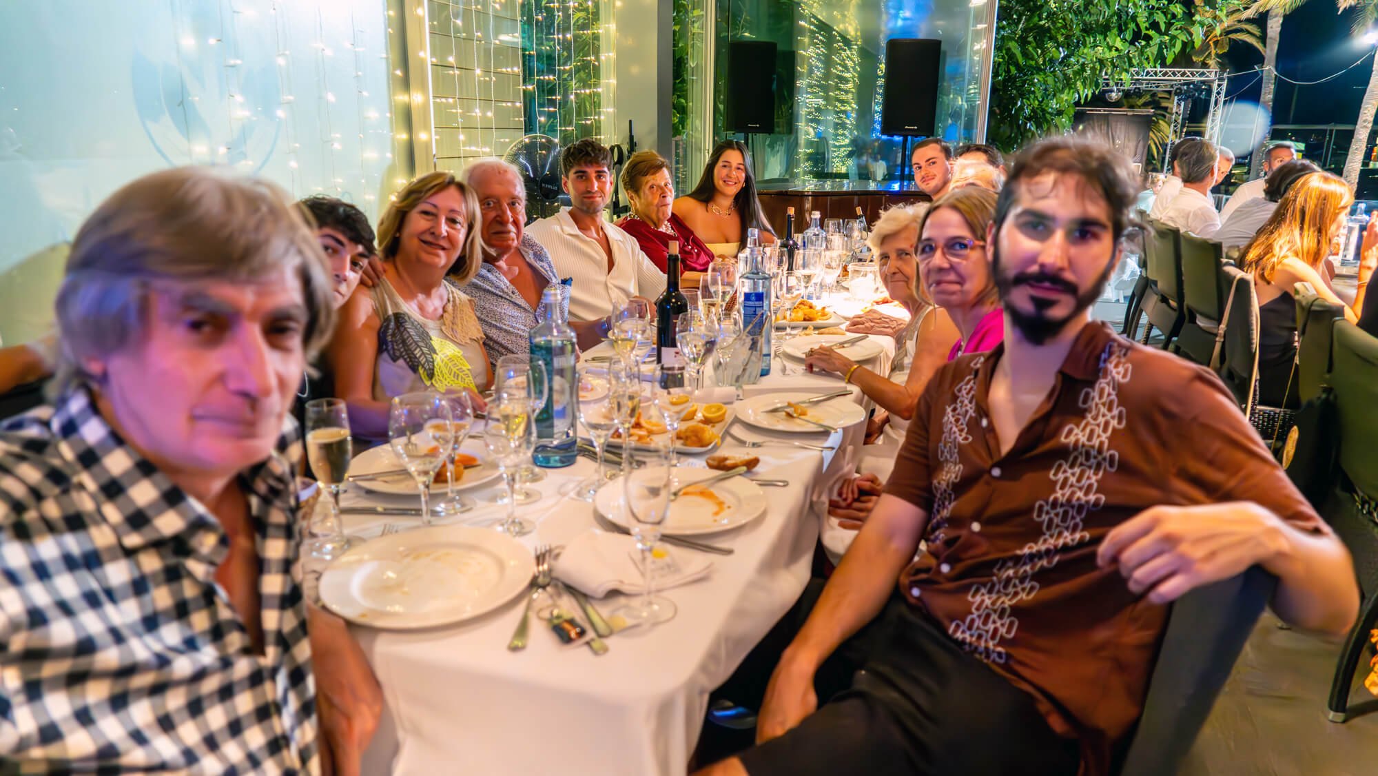 Grupo de personas sentadas en una mesa en una celebración o evento social, con decoraciones con luces y un ambiente festivo.