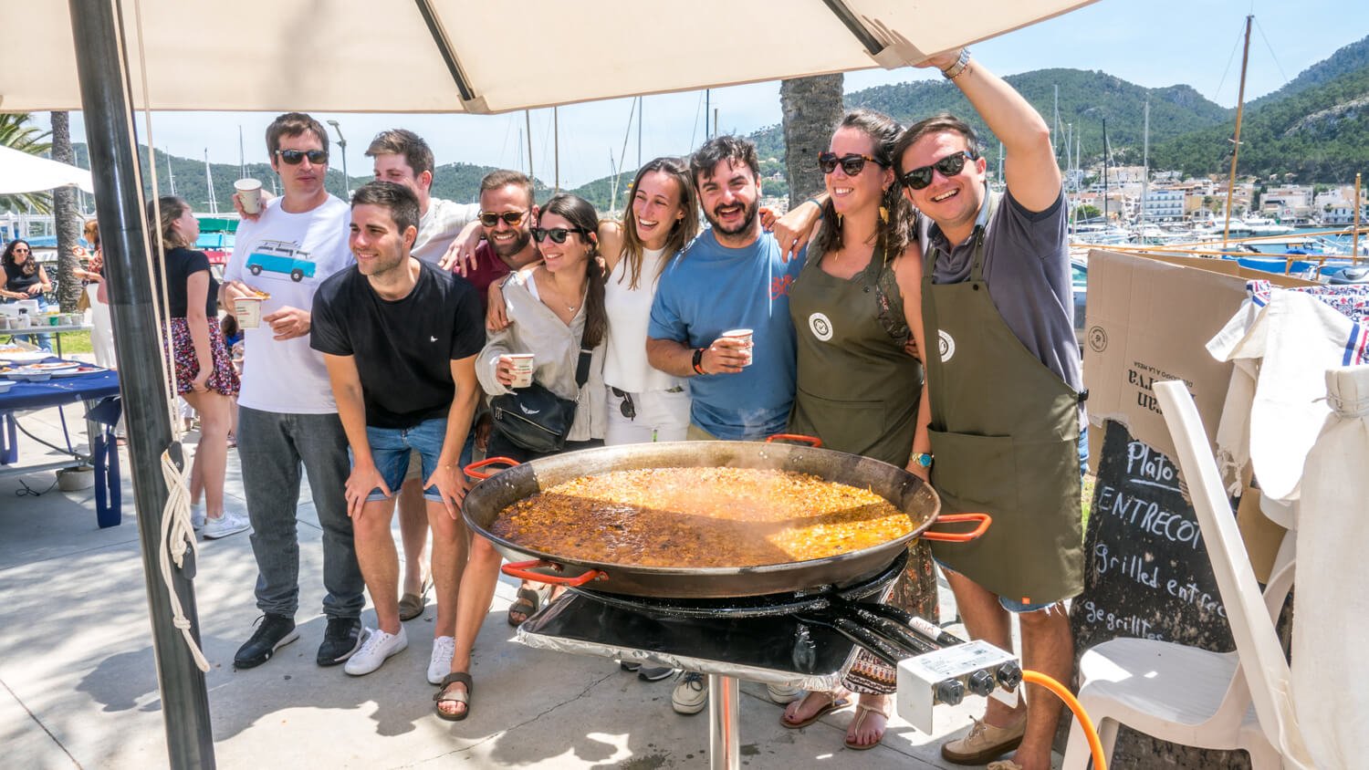 Grupo de personas sonriendo en una fiesta al aire libre con vista a un puerto con barcos, junto a una paella grande.