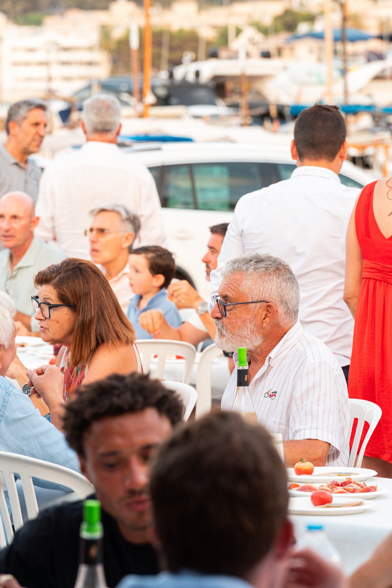 Personas en una reunión o comida al aire libre, algunas sentadas en sillas blancas, con comida en la mesa, y un fondo de barcos en un muelle.