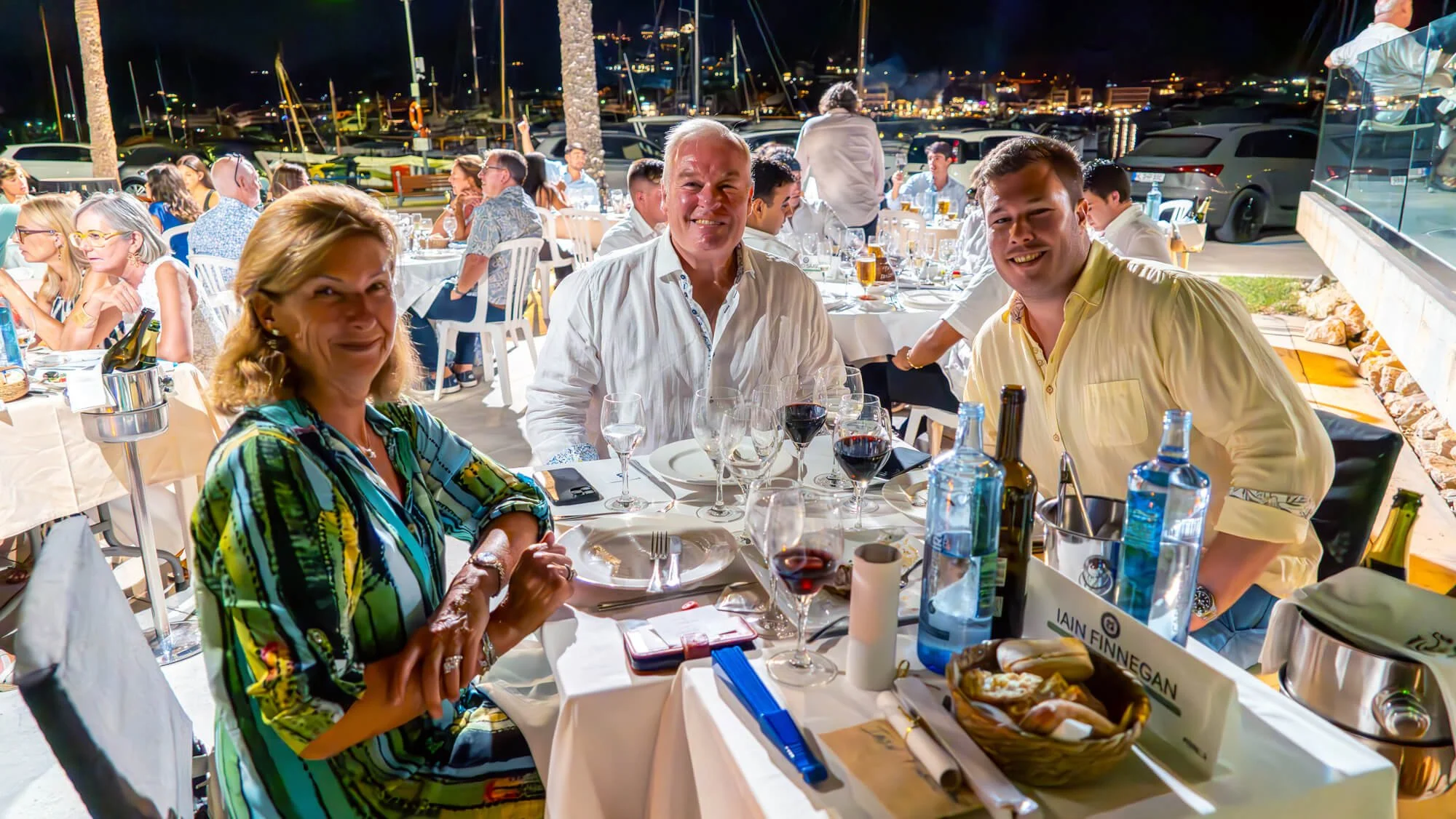 Personas en una cena al aire libre, sonriendo, con vasos de vino y comida en una mesa en un puerto con yates y luces nocturnas.