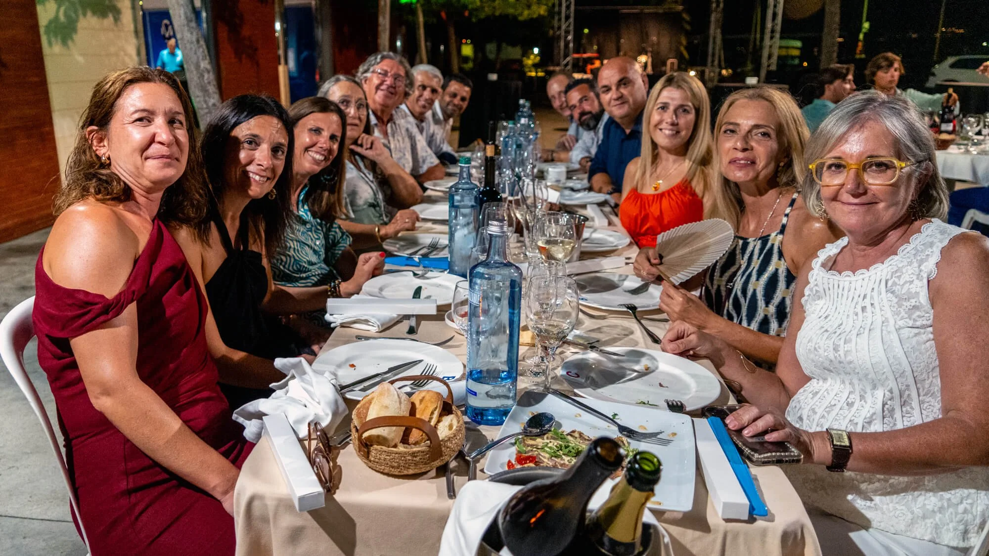 Un grupo de personas sentadas en una mesa al aire libre en una cena, con comida, bebidas y utensilios, en un ambiente nocturno.