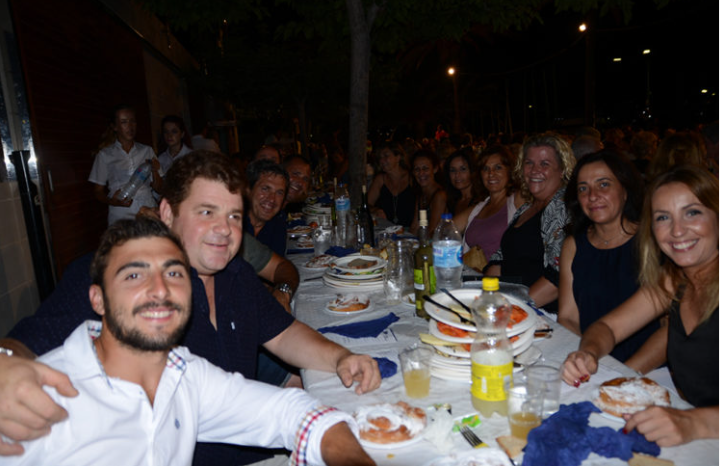 Grupo de personas sonriendo en una mesa larga al aire libre durante la noche, con comida y bebidas.