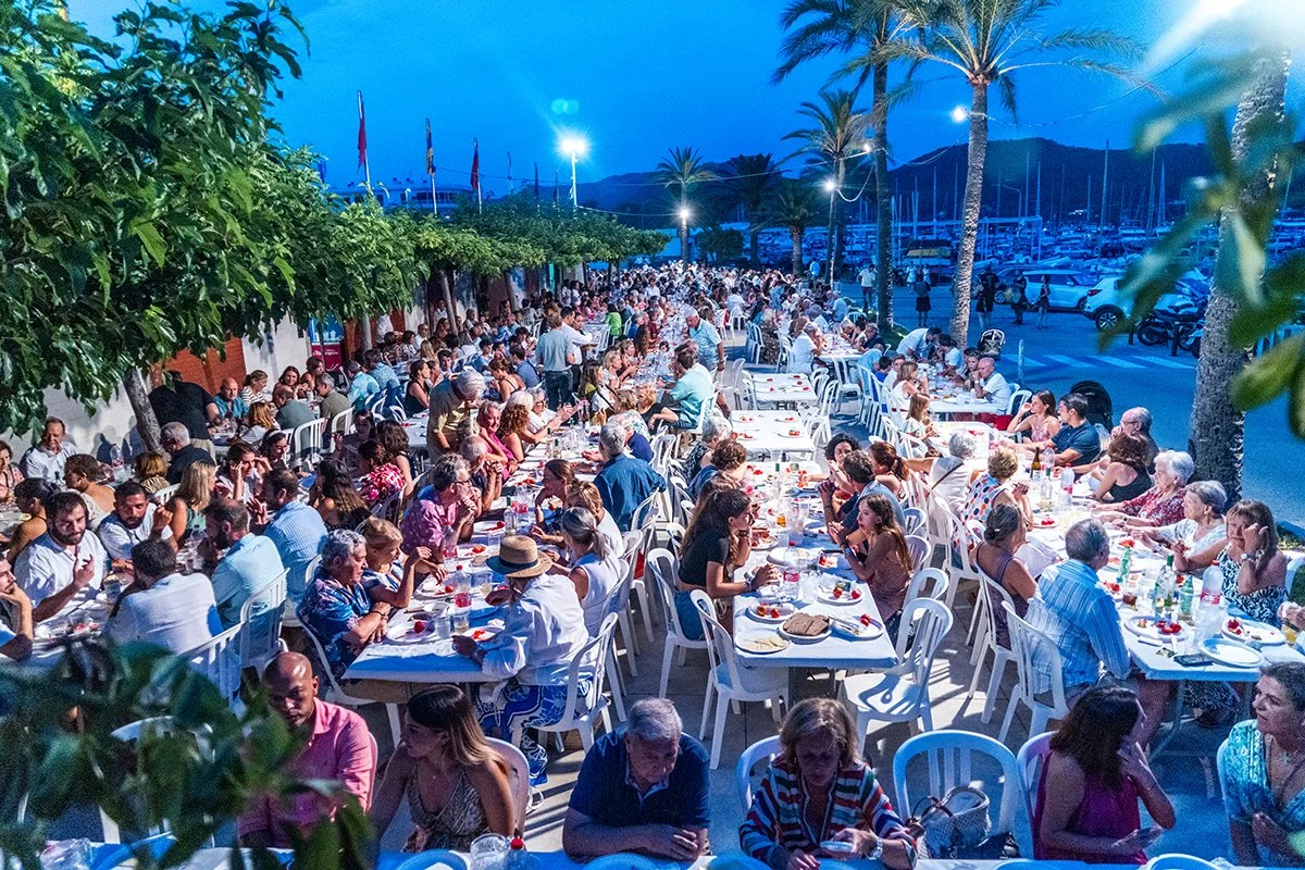 Cena al aire libre con muchas mesas llenas de personas cenando, rodeadas de palmeras y con un ambiente festivo nocturno en un puerto deportivo.
