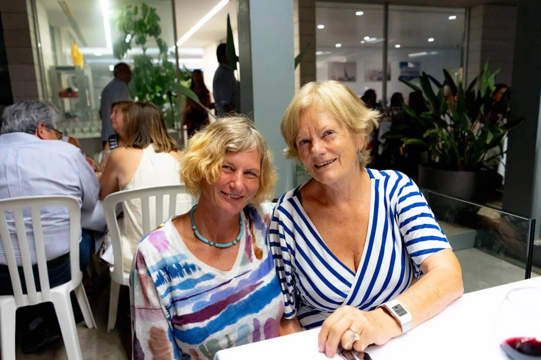 Dos mujeres sentadas en una mesa en un restaurante, sonriendo a la cámara. En el fondo, otras personas están comiendo y conversando.