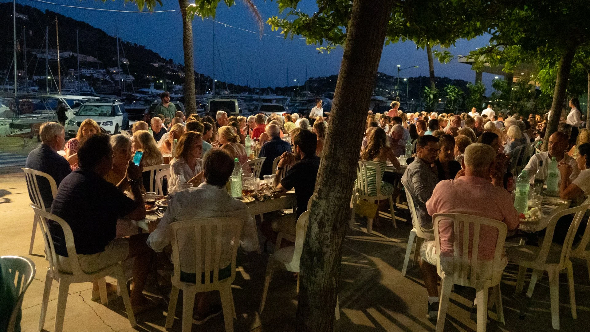 Una reunión o cena al aire libre en un puerto con muchas personas sentadas en mesas. Hay árboles y yates en el fondo, es de noche y la escena está iluminada por luces artificiales.