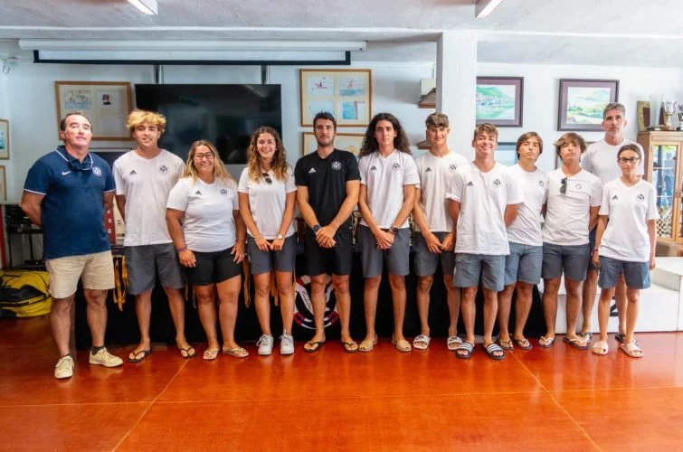 Grupo de personas, adultos y niños, posando en una sala con paredes blancas y decoraciones enmarcadas, algunas equipadas con camisetas blancas y otras con camisetas negras, en un ambiente interior.