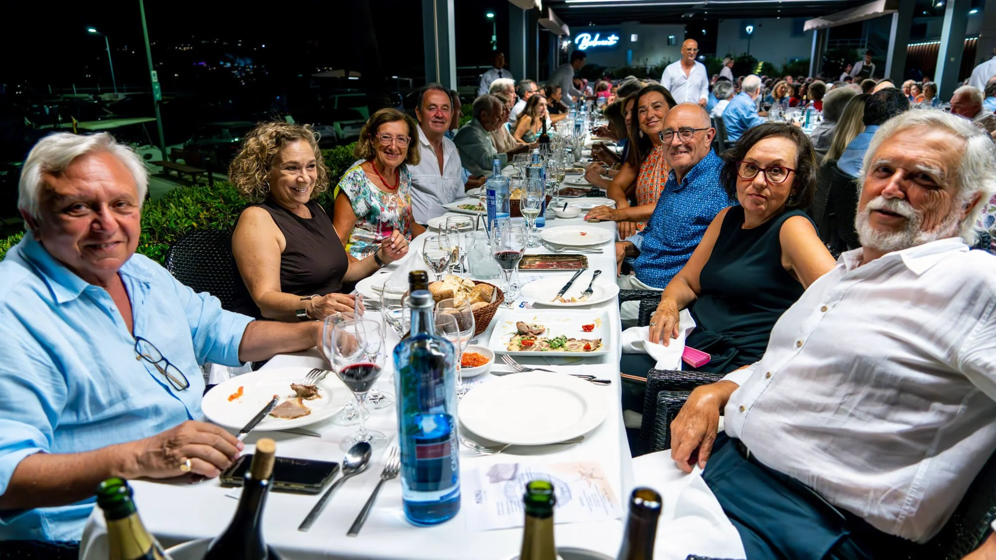 Grupo de personas sentadas en una mesa durante una cena en un restaurante, con comida y bebidas en la mesa, en un ambiente iluminado y animado.