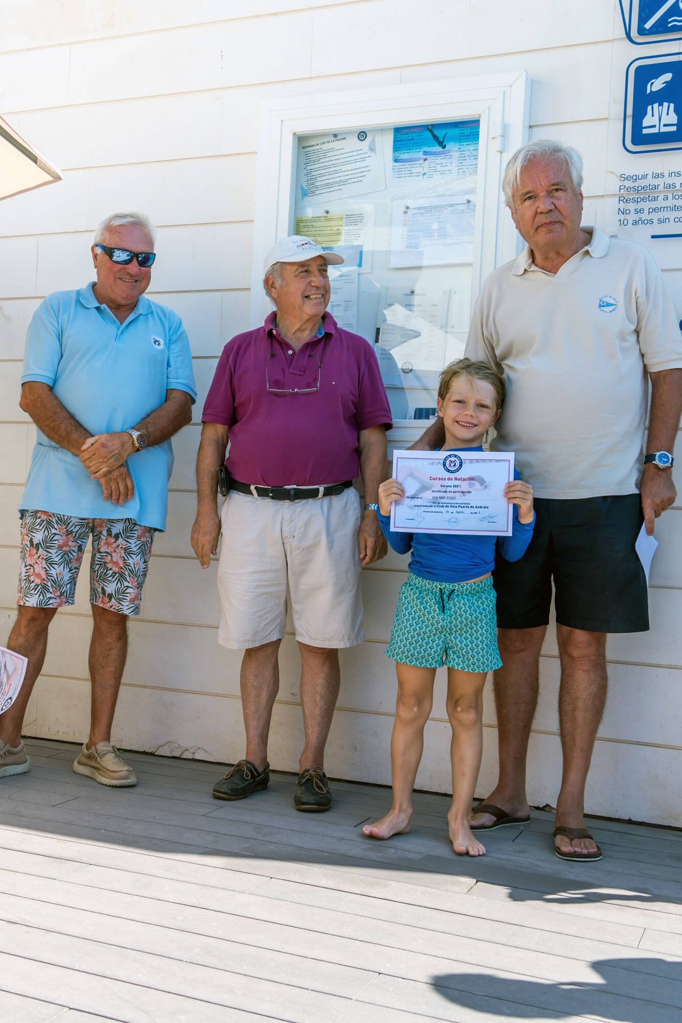 Grupo de personas, incluyendo niños y adultos mayores, posando con un diploma de natación, en un lugar exterior con pizarras y señalización.
