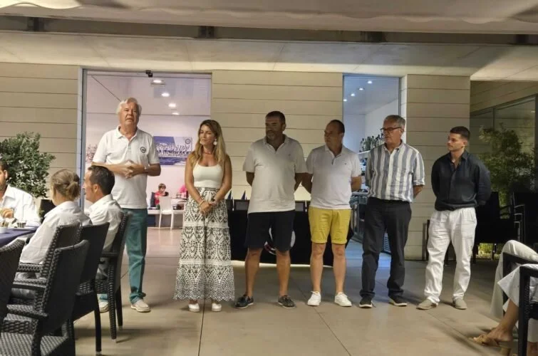Un grupo de seis personas, cinco hombres y una mujer, de pie en un espacio al aire libre, mientras una persona en el extremo izquierdo, sentado, los observa. La escena parece ser en un evento social o formal.
