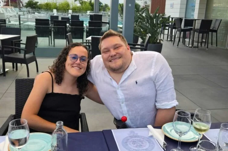 Una pareja sonriendo en una mesa de restaurante al aire libre, con vasos de agua y vino, y un menú en la mesa, en un lugar moderno y bien iluminado.
