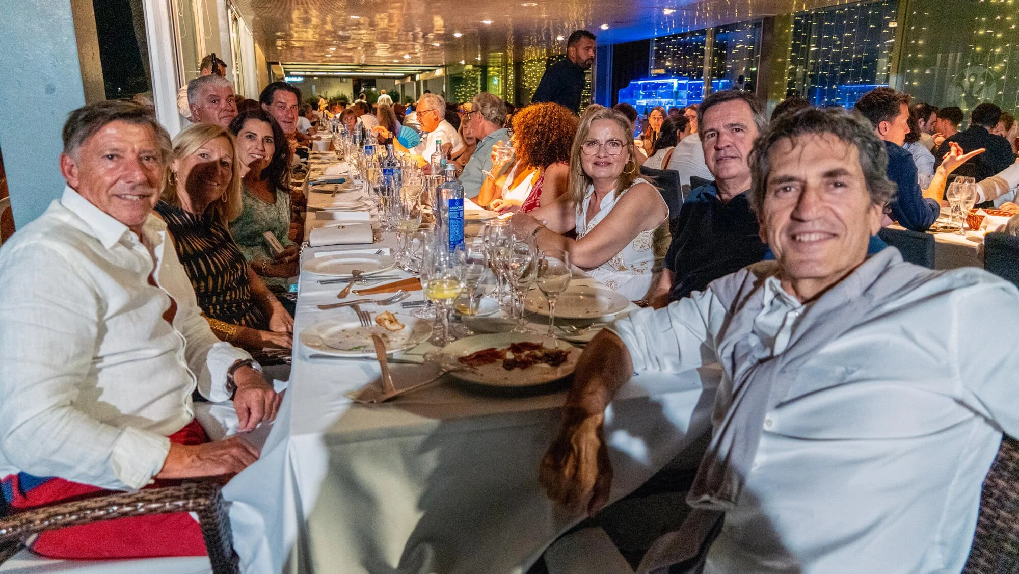 Personas en una celebración en un restaurante, sentadas en una mesa larga con comida y bebidas, en un ambiente con luces decorativas y paredes de cristal.