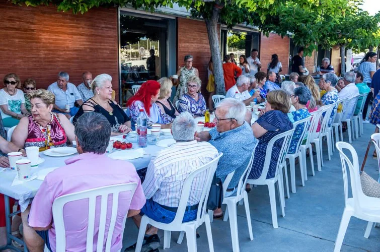 Grupo de personas mayores sentadas en una mesa al aire libre, disfrutando de una comida en un entorno exterior con árboles y un edificio de fondo.