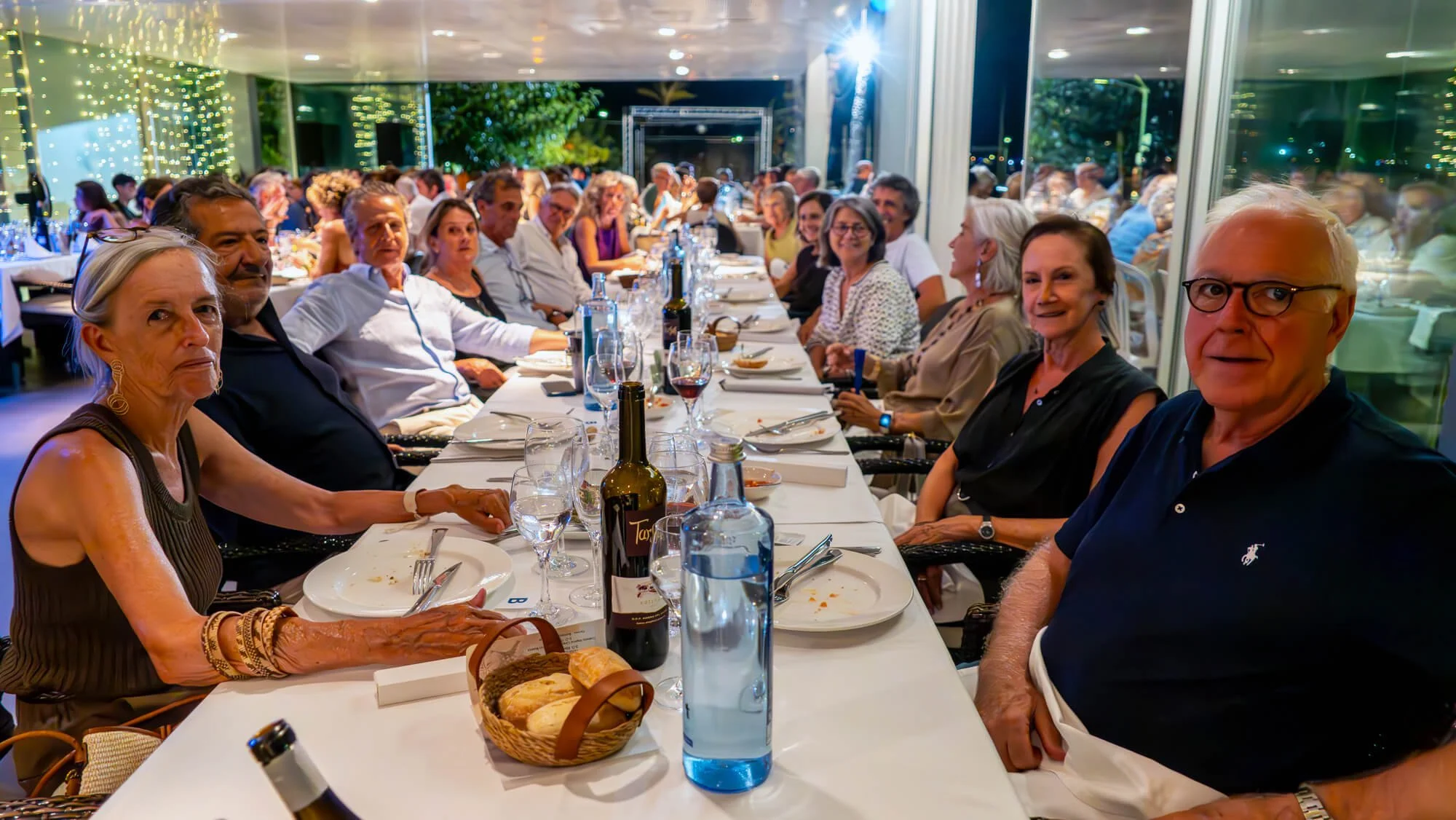 Un grupo de personas de distintas edades sentadas a lo largo de una mesa larga en un restaurante con decoración moderna y luces de fondo, disfrutando de una cena juntos.