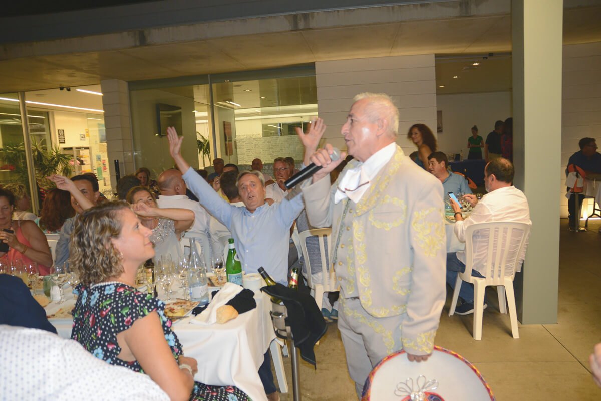 Hombre con traje claro y detalles dorados cantando con micrófono en una celebración, rodeado de personas sentadas en mesas con comida y bebidas.