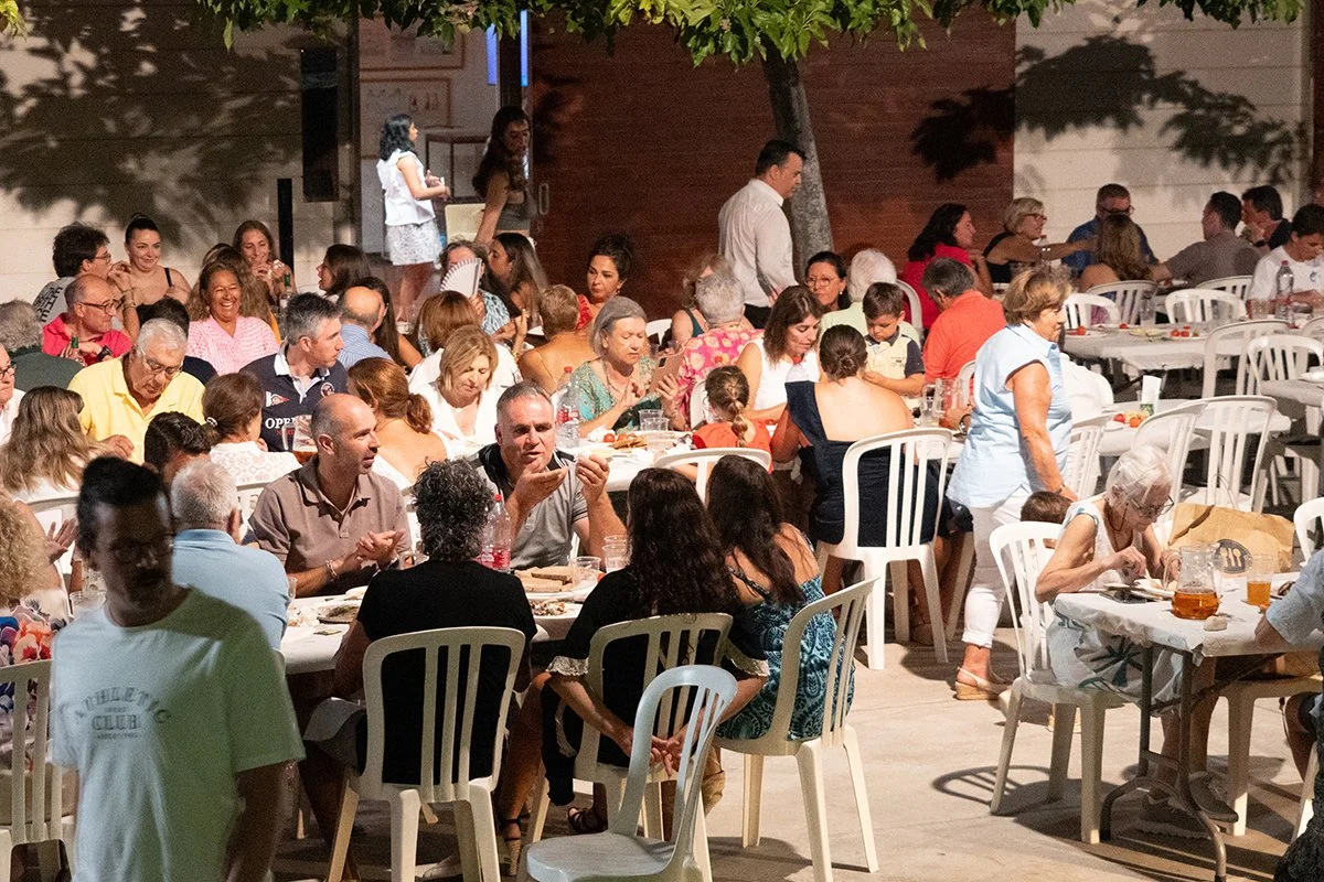Muchas personas sentadas en mesas al aire libre, disfrutando de una comida en un evento social por la noche.