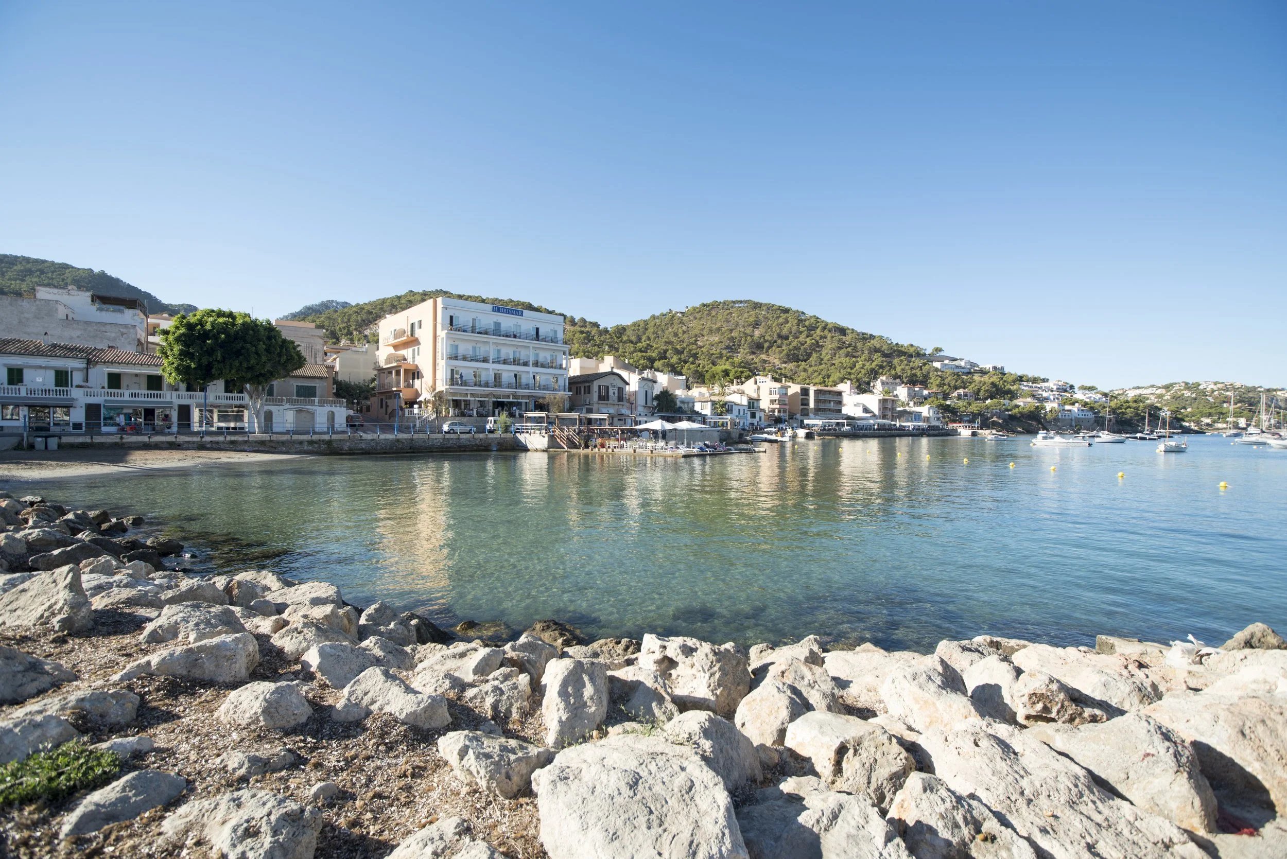 Vista de un pueblo costero con edificios blancos y un muelle, rodeado de colinas verdes, junto a un río o mar con barcos de vela y un muelle para barcos pequeños.