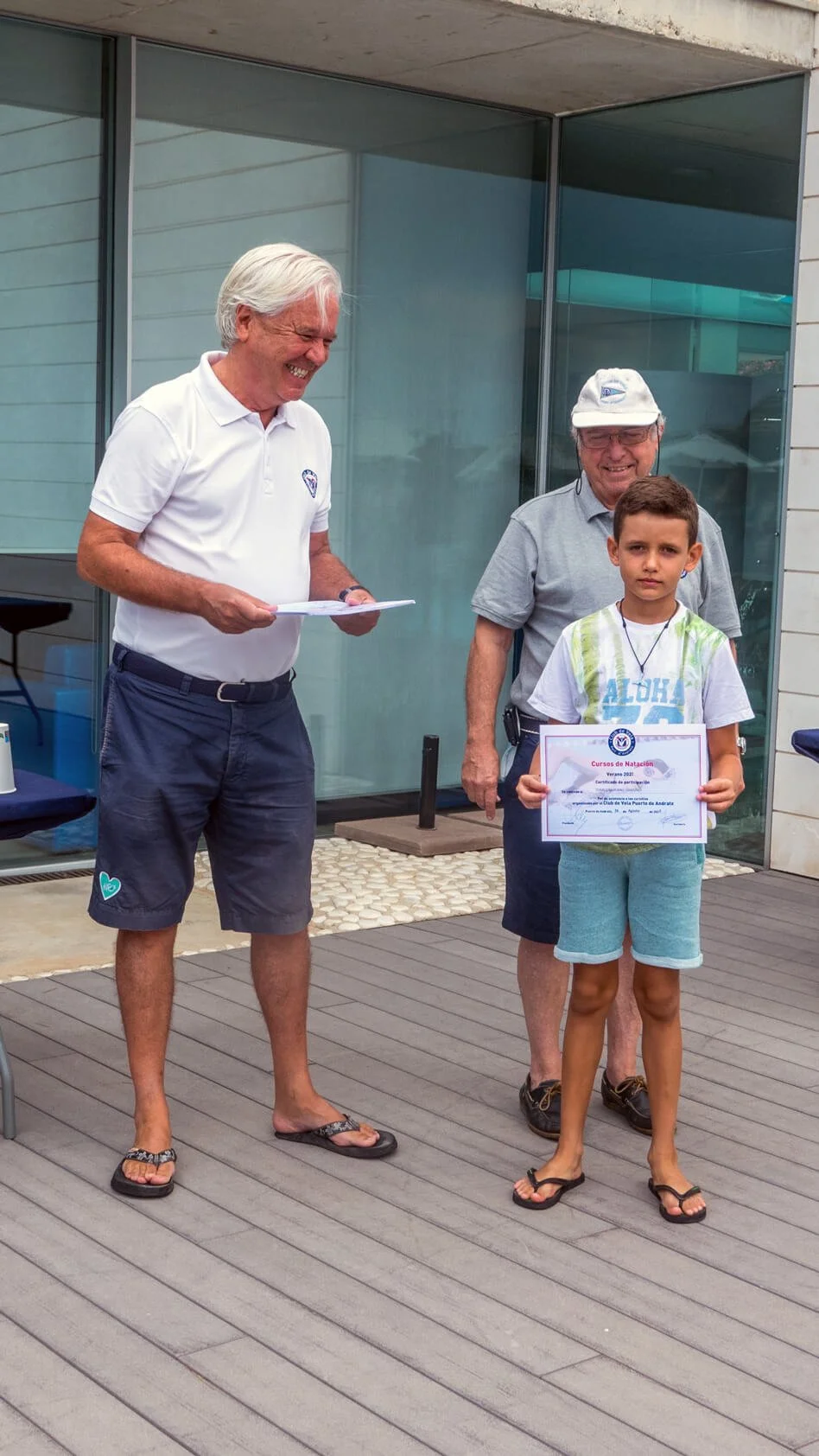 Un niño recibe un certificado de natación en una ceremonia al aire libre, acompañado por dos hombres mayores que le entregan el premio, todos son sonriendo y vestidas casualmente, en un entorno moderno con paredes de vidrio.