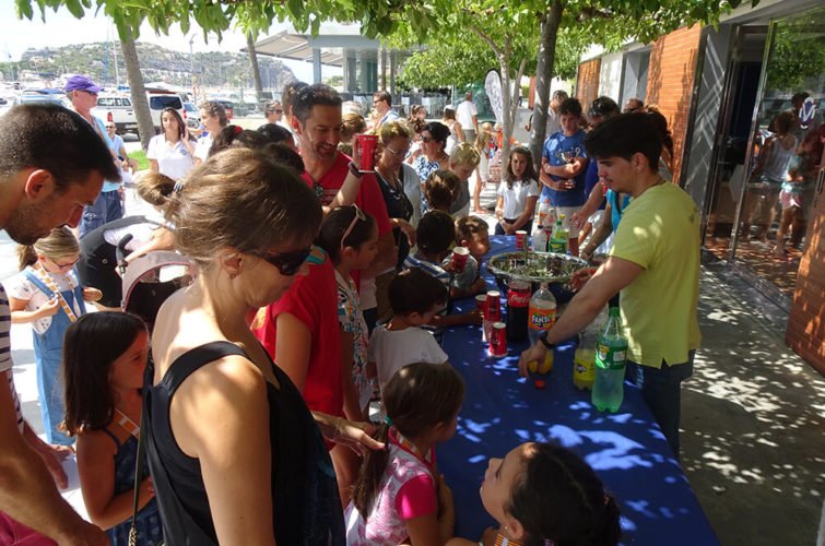 Grupo de niños y adultos en una mesa con bebidas y comida, participando en un evento al aire libre.