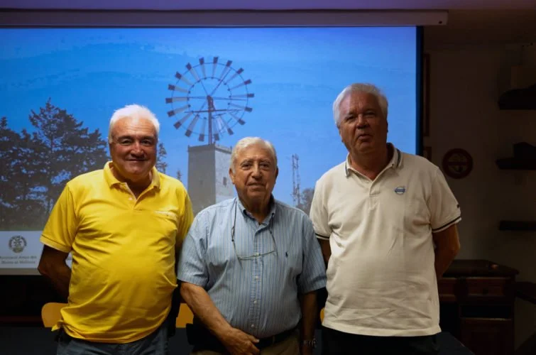Tres hombres de edad avanzada posando en una sala de conferencias con una pantalla grande detrás, en la que se proyecta una imagen de un molinillo de viento y un cielo azul.