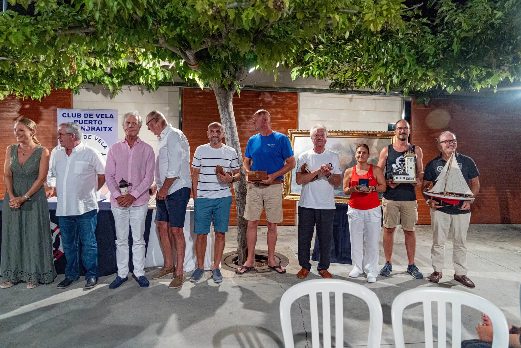 Grupo de personas en un evento nocturno en un patio, algunos sosteniendo trofeos y objetos decorativos, con un árbol y una obra de arte en el fondo, y un cartel que dice 'Club de Vela Puerto Ondraitx'.
