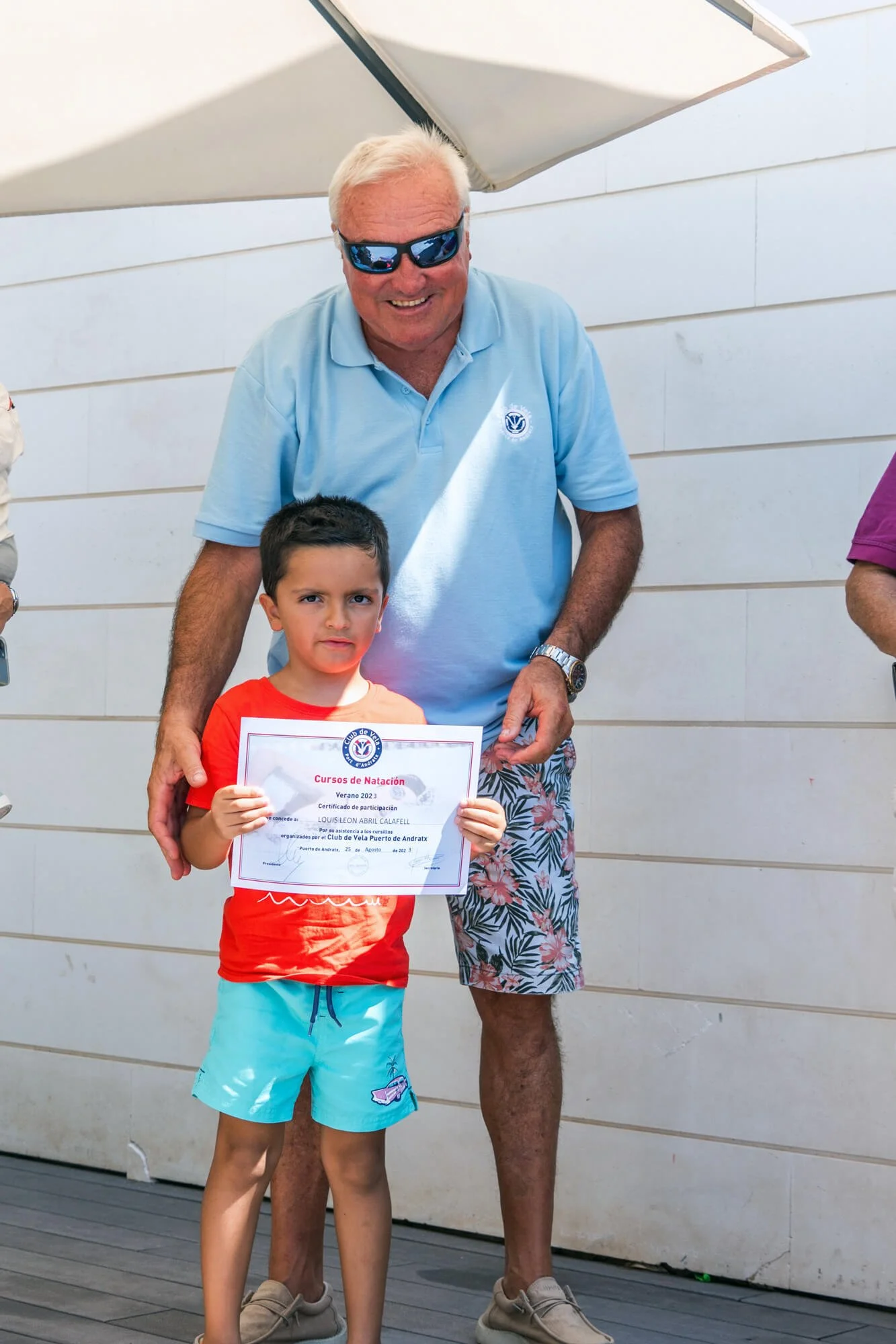 Un niño con camiseta roja y pantalones cortos azul claro recibe un certificado de natación de un hombre mayor con gafas de sol y camiseta azul claro en un evento al aire libre.