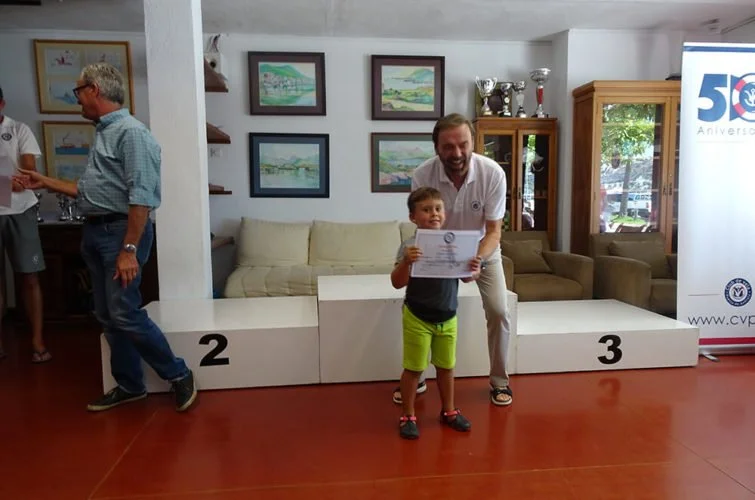 Un niño pequeño recibe un certificado de reconocimiento de un hombre sonriente en un evento de premiación en un interior decorado con cuadros y trofeos, con un podio de premios y una bandera con el logotipo del evento.