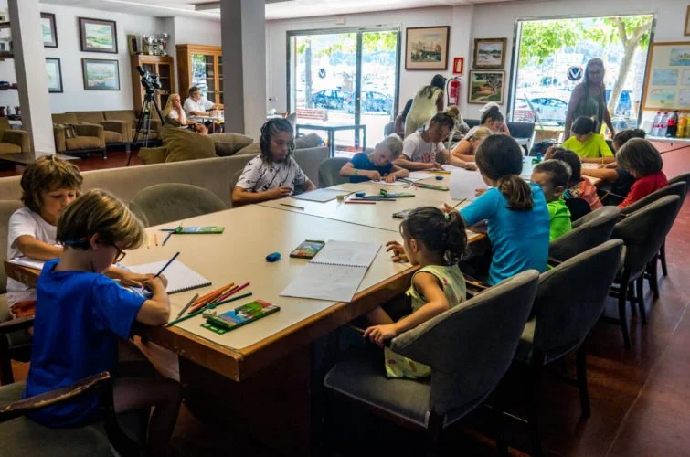 Niños sentados en una mesa en un aula, dibujando y coloreando, mientras una adulta observa en el fondo.