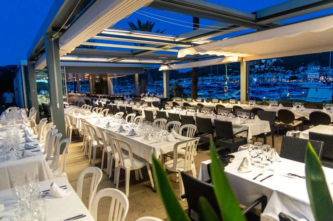 Terraza de restaurante con mesas arregladas y vista al muelle con barcos en la noche