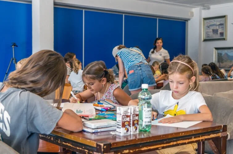 Niños estudiando en un salón, algunos escribiendo, en un ambiente escolar con paredes azules y pinturas en las paredes.