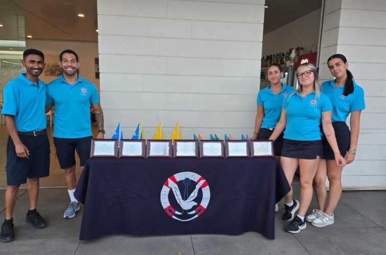 Grupo de cinco personas con camisetas azules de pie detrás de una mesa con varios premios y banderas pequeñas. La mesa tiene un mantel con un logotipo de un ancla. Están en un entorno exterior.