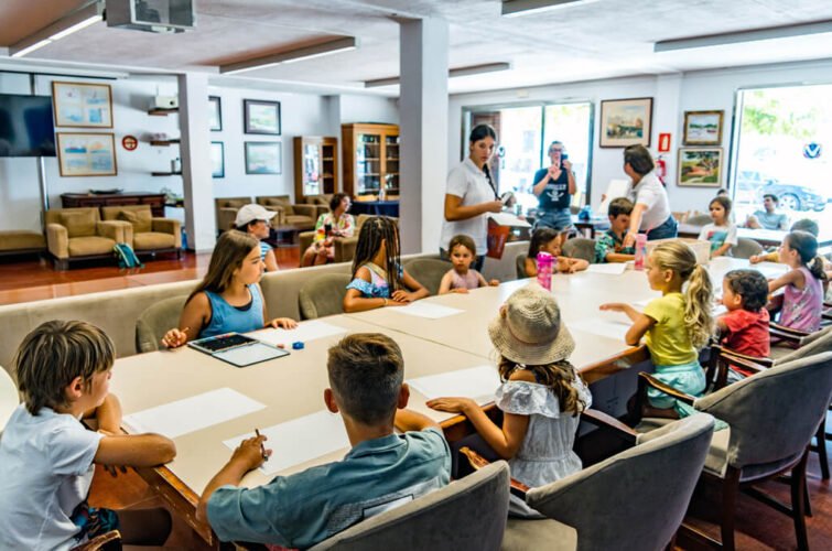 Niños en una sala de clases o taller, algunos tomando notas o pintando, con dos adultos supervisando, en un espacio luminoso con ventanas y cuadros en las paredes.