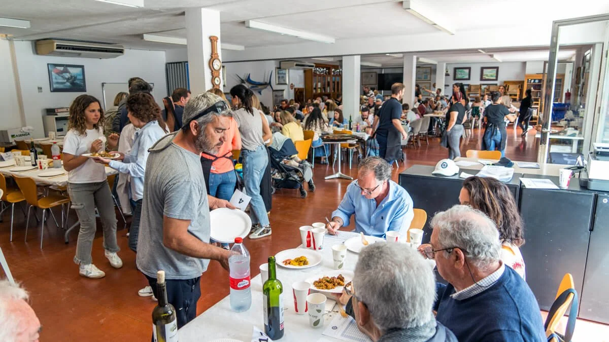 Personas disfrutando de una comida en un comedor comunitario con muchas mesas y gente sirviéndose