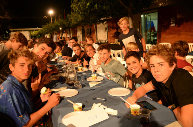 Grupo de niños y adultos sentados en una mesa durante una fiesta o reunión en exteriores por la noche.