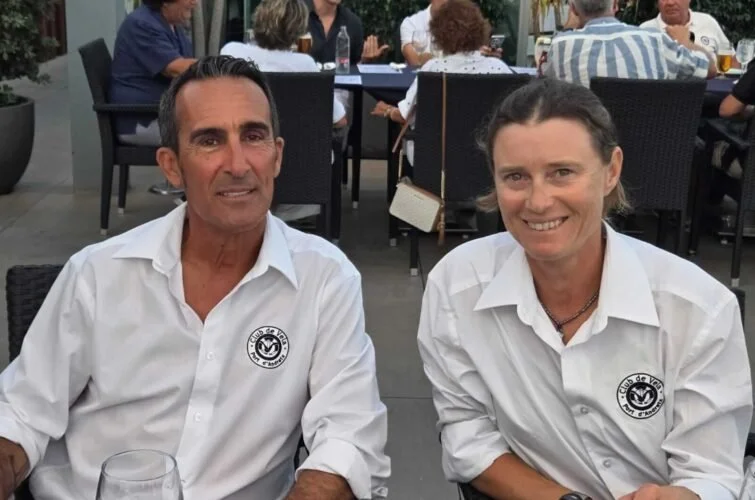 Dos personas sentadas en una mesa al aire libre, luciendo camisetas blancas con el logotipo del Colegio de Abogados, en un evento social.