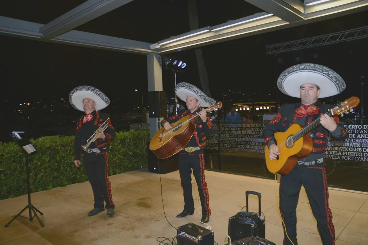 Músicos tocando en un escenario, con sombreros grandes y trajes tradicionales color negro y rojo, en una presentación nocturna.