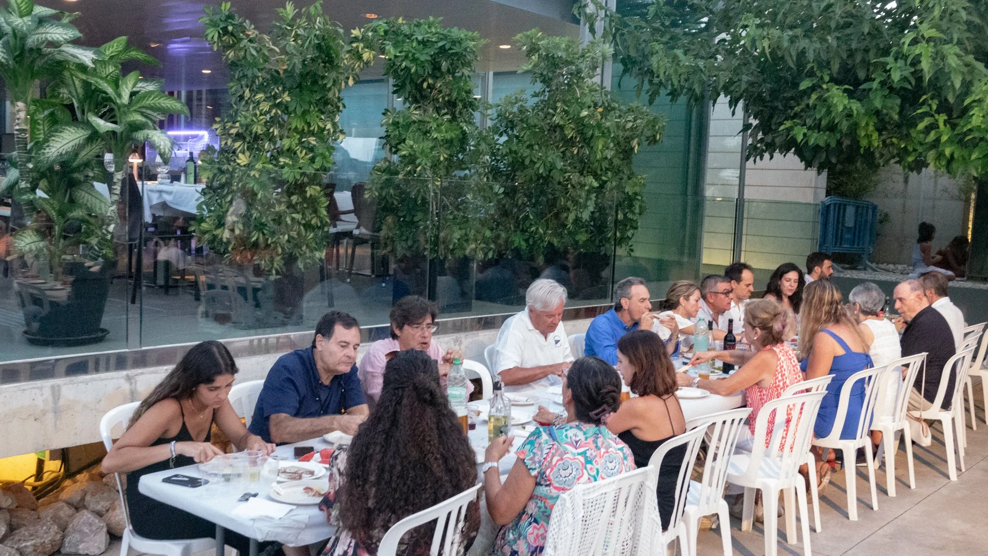 Grupo de personas sentadas en una mesa en un restaurante, comiendo y conversando, rodeado de plantas y en un ambiente semi abierto.