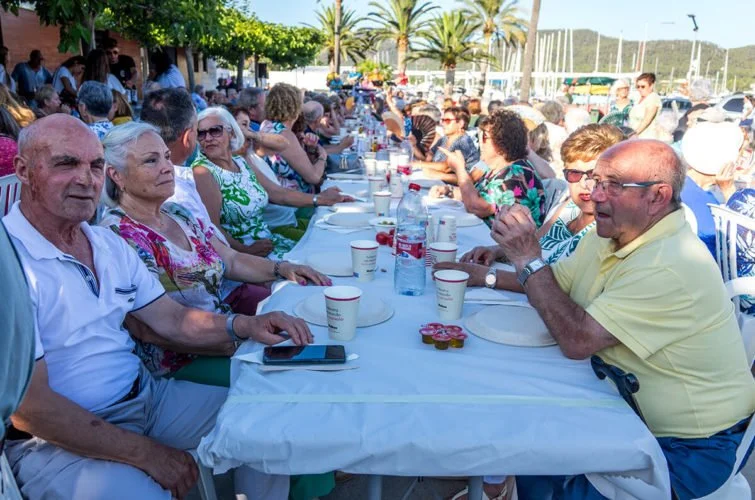 Grupo de personas mayores sentadas en una mesa larga al aire libre, disfrutando de una comida en un evento social, con fondos de palmeras y gente caminando.