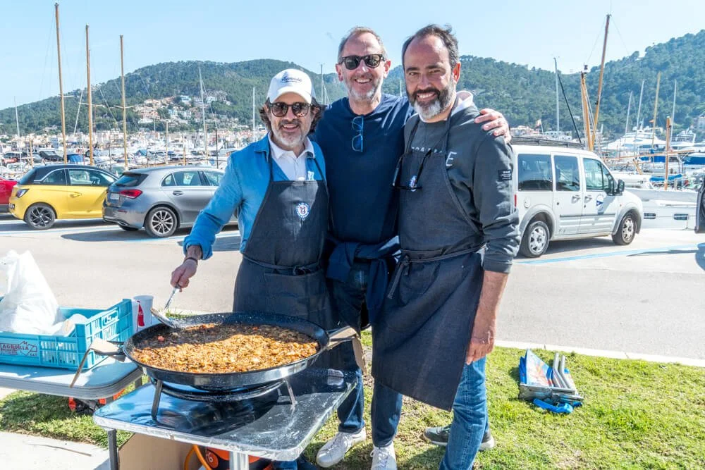 Tres hombres sonrientes en un muelle, uno cocinando en una sartén grande, con yates y colinas de fondo.