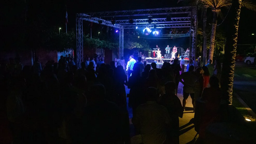 Concierto al aire libre en la noche con público disfrutando de la música en un escenario iluminado con luces azules y decorado con palmeras.