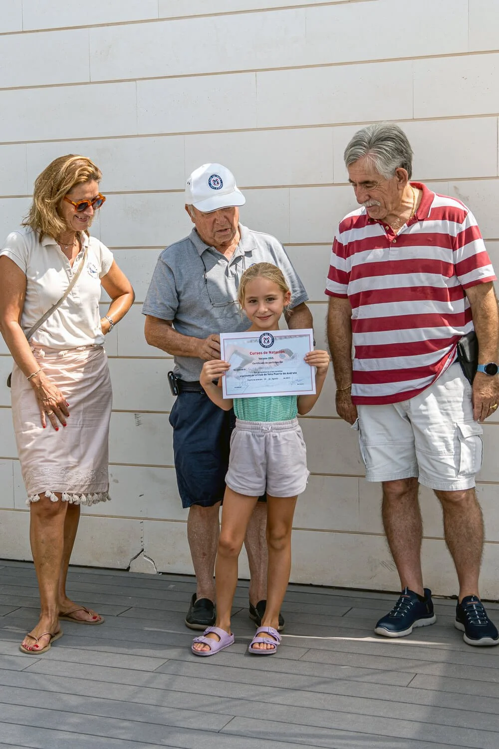 Niña recibiendo certificado de natación en una ceremonia al aire libre, acompañada por varias personas adultas.